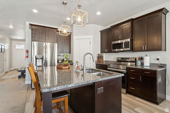 Kitchen with appliances with stainless steel finishes, dark brown cabinetry, dark stone counters, an island with sink, and hanging light fixtures | Image 16