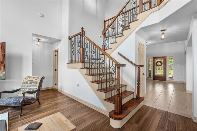 This inviting home features a grand entrance with a beautiful staircase and decorative iron railings. The open layout includes a cozy sitting area with wood flooring, leading to a bright foyer with tiled flooring and a welcoming front door that offers plenty of natural light. | Image 22