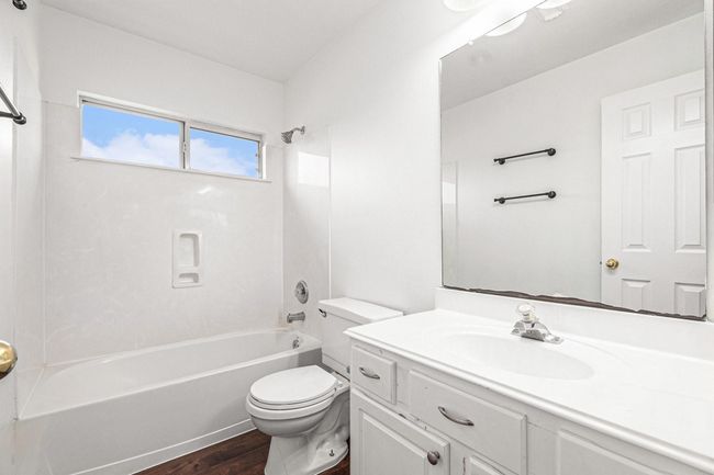This bathroom features a clean, modern design with a white vanity, large mirror, and a bathtub with a shower. The space is bright due to a window that allows natural light to flood in. | Image 16