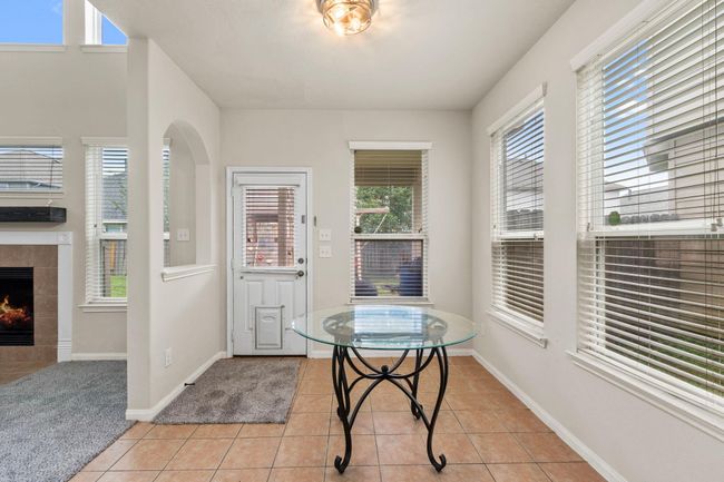 This photo showcases a bright, cozy space with tile flooring and ample natural light from multiple windows. It features a small glass table and a doorway leading to the outdoors, with a view of a fireplace in the adjacent carpeted room. | Image 12