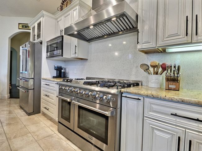 Kitchen featuring wall chimney exhaust hood, appliances with stainless steel finishes, arched walkways, white cabinetry, and decorative backsplash | Image 15