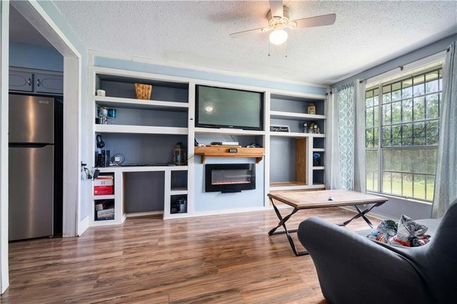 Living area with a electric fireplace, wood finished floors, ceiling fan, a textured ceiling, and built in shelves | Image 5