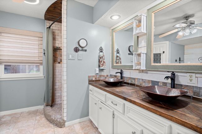 This primary bathroom features a rustic design with a double vanity topped by stylish vessel sinks and framed mirrors. The walk-in shower has a curved entry with stone accents, with lots of natural light filtering in! | Image 25