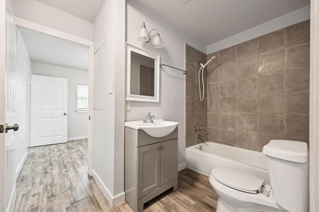 This bathroom features a modern design with neutral-toned tiles around the bathtub and shower. It includes a compact vanity with a mirror and updated fixtures. The flooring extends into an adjacent room, providing a cohesive look. | Image 8