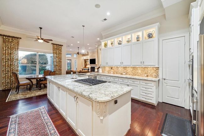 This kitchen features elegant white cabinetry with granite countertops, a spacious island with a cooktop, and dark hardwood flooring. The open layout connects to a cozy dining area and living space with large windows and a fireplace, creating a bright and welcoming atmosphere. | Image 12