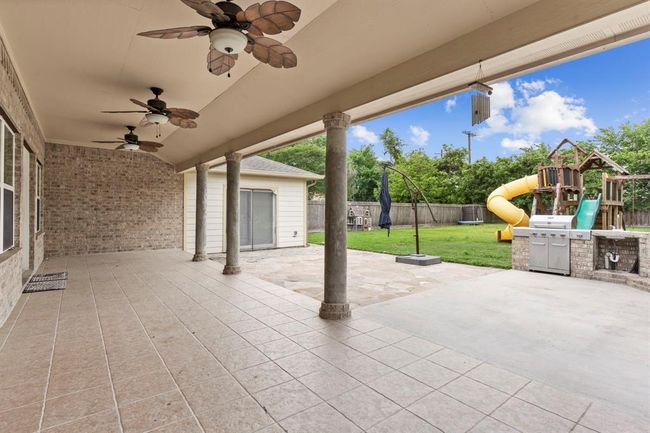 Spacious covered patio with ceiling fans, tile flooring, and an outdoor kitchen, overlooking a fenced backyard with a playset. Ideal for entertaining and family activities. | Image 40