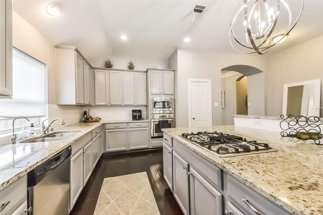 Kitchen features sleek granite countertops, a central island with a gas cooktop, and light gray cabinetry. It includes stainless steel appliances, dark wood flooring, and modern lighting, creating a bright and inviting space. | Image 11