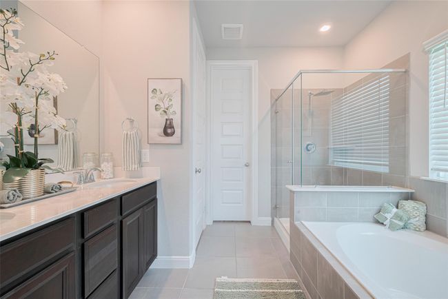 This bathroom features a modern design with a spacious glass-enclosed shower, a separate bathtub, and a dual-sink vanity with dark cabinetry. It has neutral tile flooring, a large mirror, and ample natural light from the window with blinds. | Image 26