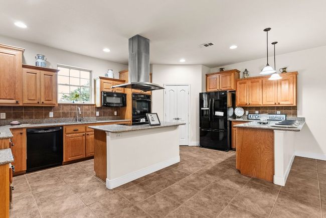 Kitchen with tasteful backsplash, decorative light fixtures, black appliances, island range hood, and light stone counters | Image 11