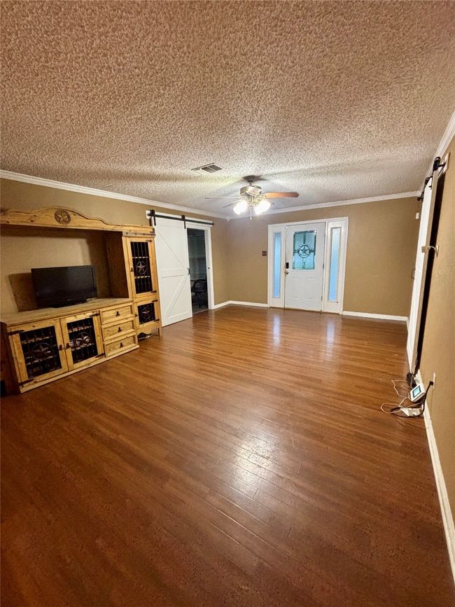 Unfurnished living room featuring a barn door, ornamental molding, wood finished floors, a textured ceiling, and ceiling fan | Image 5