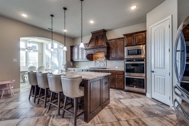 Kitchen featuring appliances with stainless steel finishes, decorative light fixtures, custom exhaust hood, a kitchen island with sink, and light stone counters | Image 12