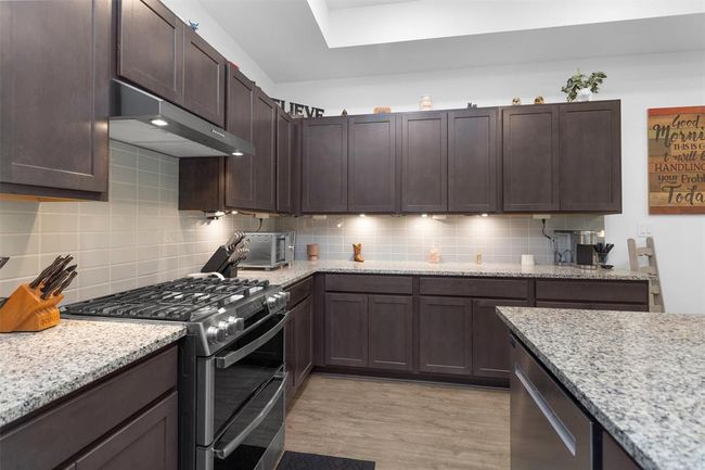 This show-stopping kitchen is the true heart of the home—designed for both everyday ease and upscale entertaining. Featuring an oversized granite island with bar seating, a custom designer vent hood, stainless steel appliances, and stylish tile backsplash, it’s as beautiful as it is functional. The massive walk-in pantry offers storage galore, keeping everything tucked away yet within reach. | Image 18