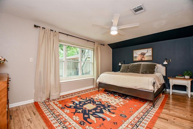 This cozy bedroom features a large window allowing natural light, a modern ceiling fan, and a stylish accent wall. The room is complemented by a vibrant patterned rug and hardwood flooring, creating a warm and inviting atmosphere. | Image 25