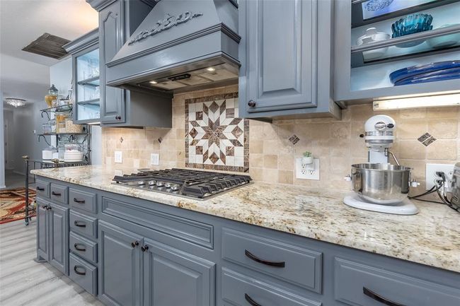 Kitchen with stainless steel gas stovetop, decorative backsplash, light wood-type flooring, custom range hood, and light stone counters | Image 5