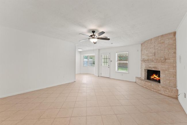 This spacious living area features a cozy corner fireplace with brick detail, tiled flooring, and ample natural light from multiple windows. The room is enhanced by a ceiling fan and offers access to the outdoors through a door with glass panels. | Image 8