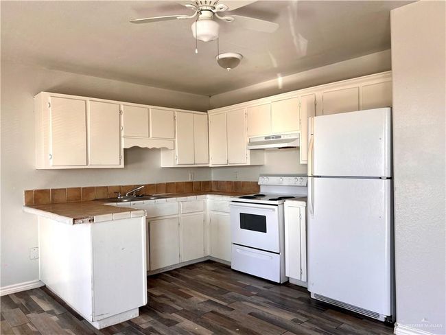 Kitchen with ceiling fan, under cabinet range hood, white cabinets, white appliances, and dark wood-style flooring | Image 4