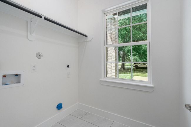 This photo shows a bright laundry room with white walls, a window providing natural light, utility hookups, and a shelf with a hanging rod. | Image 30