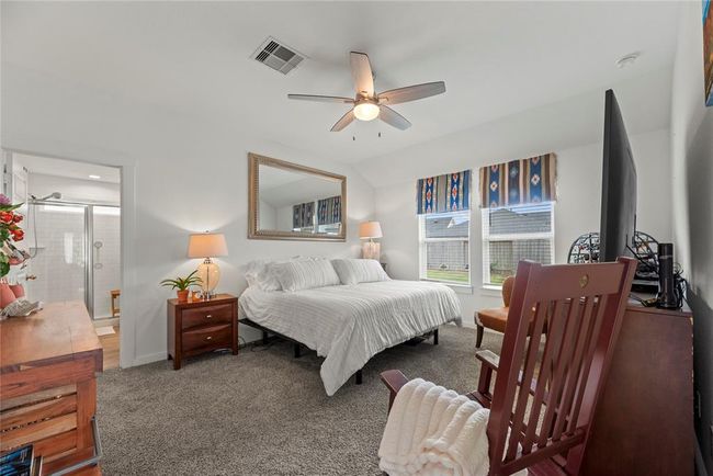 The primary suite is a serene retreat, featuring soft carpet underfoot for added comfort. Two windows bring in natural light, brightening the room and enhancing the airy feel. The light walls create a calm, neutral backdrop, while the ceiling fan ensures a cool, comfortable environment. The suite also includes a connecting bathroom, offering convenience and privacy, making it the perfect space for relaxation. | Image 10