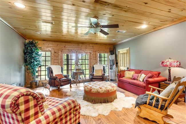This cozy living room features warm wood paneling on the ceiling, hardwood floors, and large windows that let in plenty of natural light. The space is comfortably furnished with a mix of upholstered chairs, a plush sofa, and a central ottoman, creating an inviting atmosphere. A ceiling fan and decorative lighting add to the room's charm. | Image 29