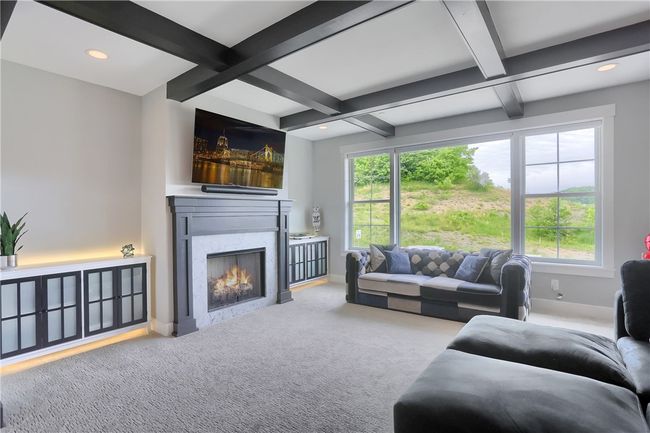 This sophisticated family room features a coffered ceiling, built-in lighted cabinetry, and a dramatic fireplace with marble surround—framed by oversized windows that flood the space with natural light and lush views, all seamlessly connected to the designer kitchen. | Image 20