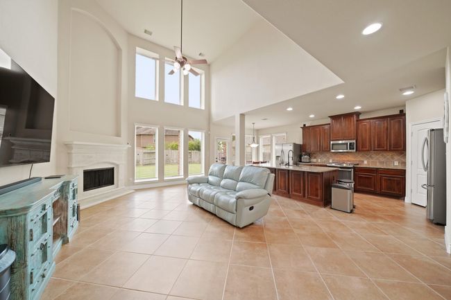 This spacious and bright open-concept living area features high ceilings, large windows, and a modern kitchen with dark wood cabinetry and stainless steel appliances. The room includes a cozy fireplace and tiled flooring, seamlessly connecting to the dining space, perfect for entertaining and family gatherings. | Image 11