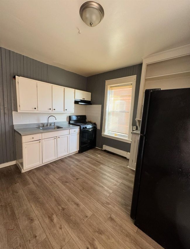 Kitchen with wood finished floors, a sink, a baseboard heating unit, black appliances, and white cabinetry | Image 7