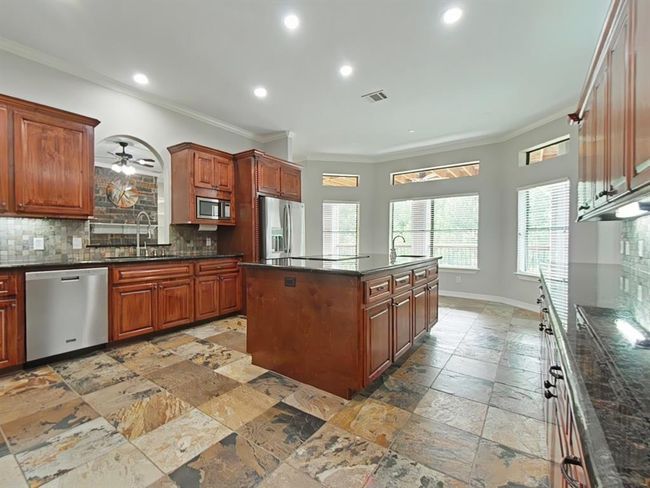 Spacious kitchen with dark granite countertops and rich wooden cabinetry. Features an island with a cooktop, stainless steel appliances and slate tile flooring. | Image 7