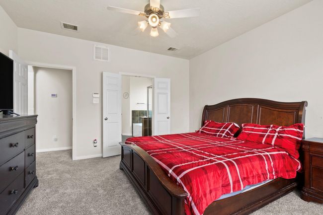 This spacious bedroom features a plush carpet, a ceiling fan, and elegant dark wood furniture. The room includes a large bed with vibrant red bedding and offers access to an en-suite bathroom through double doors. | Image 16
