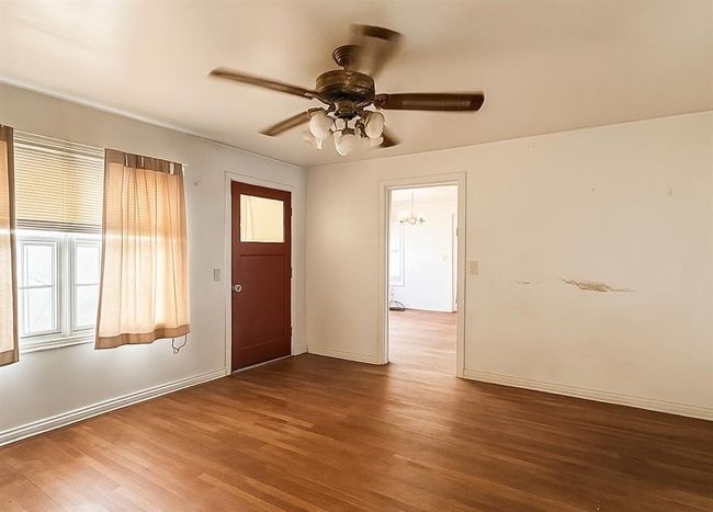 This room features hardwood flooring, a ceiling fan, and a red door with a window. Natural light filters in through two windows with light curtains. The space leads into another room with additional lighting. | Image 5