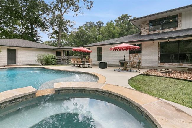 This photo showcases a spacious backyard with a large swimming pool and a hot tub, surrounded by a patio area featuring outdoor seating with umbrellas. The house exterior is brick with ample windows, set against a backdrop of lush trees, providing a private and relaxing atmosphere. | Image 42