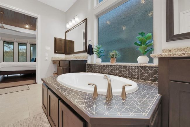 The modern bathroom with a large soaking tub, elegant tile surround, and dual sinks with granite countertops. The space is brightened by a large frosted window. | Image 29