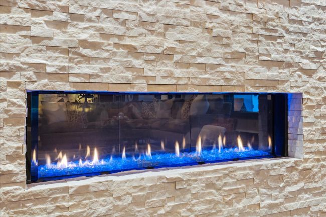 A modern, linear gas fireplace anchors the living space with mesmerizing flame effects dancing over crushed glass and a glowing blue accent. Framed by a textured stacked-stone surround in a neutral palette, the fireplace can be changed to most any color depending on the mood desired, which adds both warmth and contemporary sophistication. It's the perfect focal point for cozy evenings or upscale entertaining, seamlessly blending form and function. | Image 14