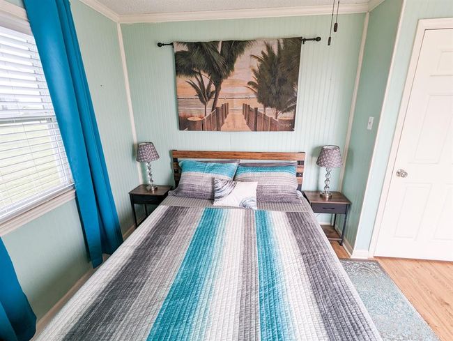 This cozy bedroom features a comfortable bed with a striped quilt, flanked by two bedside tables with lamps. The room is painted in a soothing light green, complemented by blue curtains. A beach-themed artwork adds a relaxing touch. | Image 15