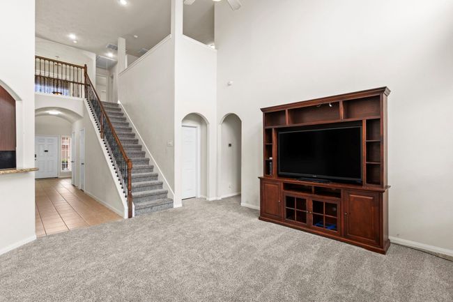 This spacious living area features plush carpeting and a tall ceiling, providing an open feel. A staircase with elegant wood railing leads to the upper level, while a large entertainment unit offers ample storage and display space. The room is bright, with neutral tones that add to the inviting atmosphere. | Image 14