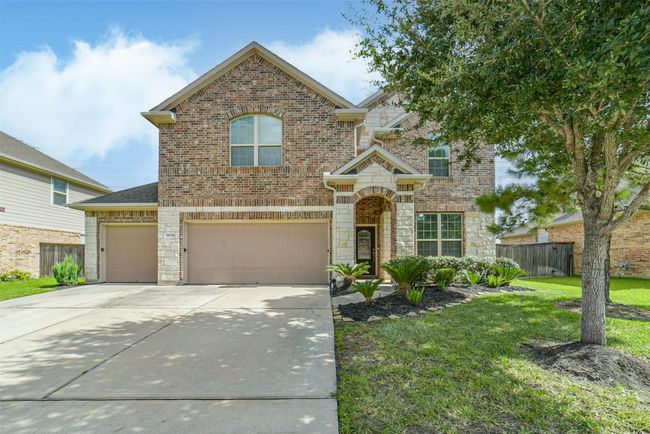 This photo showcases a two-story brick home with a three-car garage. The front yard features well-maintained landscaping with a tree and shrubs, and a clean driveway leading to the entrance. | Image 5