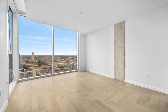 Step into this sleek and contemporary bedroom featuring floor-to-ceiling windows that frame spectacular views of the city and beyond. The natural light floods the room, enhancing the airy ambiance created by the light wood flooring and neutral color palette. The exposed concrete accent wall adds an industrial touch to the minimalist design. | Image 7