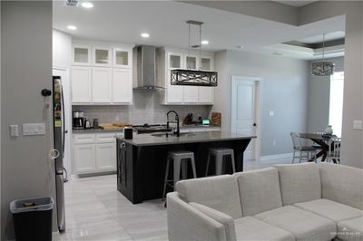 Kitchen featuring wall chimney range hood, an island with sink, white cabinetry, pendant lighting, and decorative backsplash | Image 2