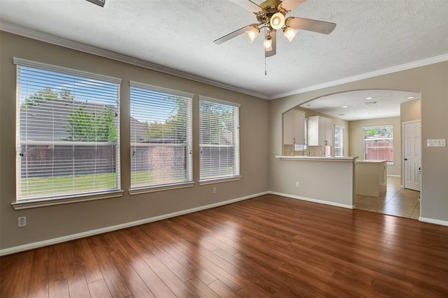 Bright and spacious living area with large windows, wood flooring, and a ceiling fan. It opens to a modern kitchen with a breakfast bar and tiled floors, overlooking a fenced backyard. | Image 17