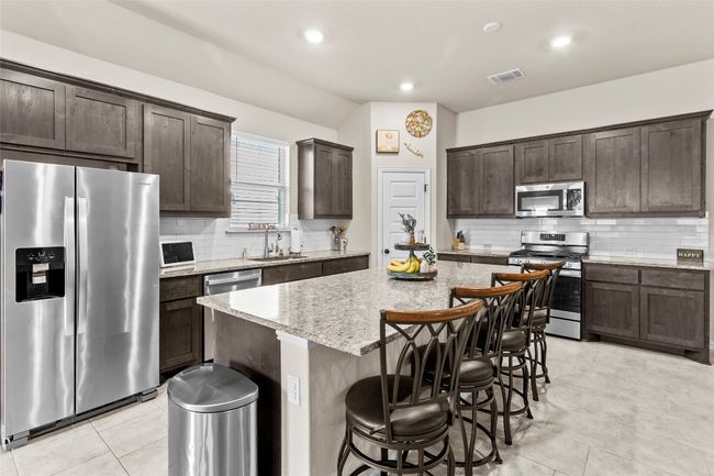 Kitchen with appliances with stainless steel finishes, a kitchen island, dark brown cabinets, decorative backsplash, and light stone countertops | Image 8