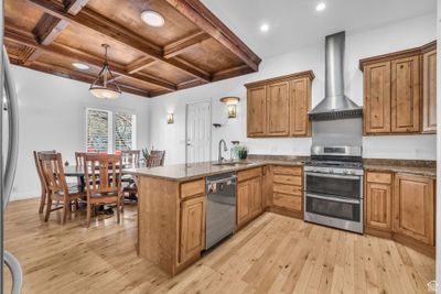 Kitchen featuring light hardwood / wood-style flooring, appliances with stainless steel finishes, wall chimney range hood, coffered ceiling, and kitchen peninsula | Image 1