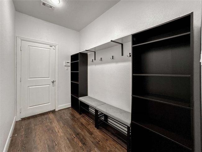 Stay organized with this mudroom with luxury vinyl flooring, white walls, and a door leading to the garage. It features black shelving units, a bench with hooks above for hanging, and overhead lighting. | Image 17