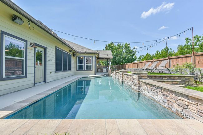 The backyard features a sleek lap pool alongside a modern house, complemented by a stone wall and cozy poolside seating. String lights add a warm, inviting touch, perfect for relaxation and entertainment. | Image 30