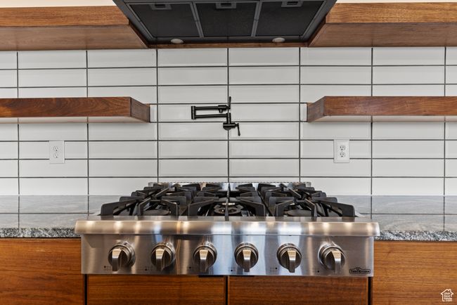 Kitchen view of stainless steel gas stovetop, ventilation hood, decorative backsplash, dark stone countertops, and pot filler | Image 8