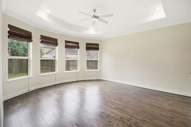This spacious room features elegant wood flooring, a tray ceiling with recessed lighting, and a modern ceiling fan. Large windows with wooden blinds offer ample natural light and views of the fenced yard, creating a bright and inviting atmosphere. The bay window adds additional seating space. | Image 13