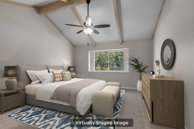 This is a virtually staged primary bedroom with a vaulted ceiling and exposed wooden beams. It features a cozy bed with neutral bedding, a ceiling fan, a dresser, a large window allowing natural light, and decorative elements like a mirror and a plant for a modern, inviting look. | Image 24