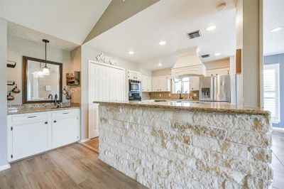 This bright, modern kitchen features white cabinetry, granite countertops, and stainless steel appliances. It has a stone breakfast bar, adding warmth and texture. The space is well-lit with natural light and recessed lighting. | Image 3