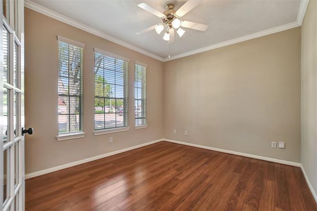 This room features hardwood flooring, neutral walls, and abundant natural light from three large windows. It includes a ceiling fan and French doors, making it ideal for a home office or cozy living space. | Image 8