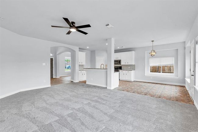 This spacious open-plan living area features new carpeting, a modern kitchen with white cabinetry, stainless steel appliances, and a breakfast bar. The room is well-lit with natural light from large windows, and includes a ceiling fan and tile flooring in the dining area. | Image 10