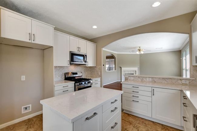 This kitchen features modern white cabinetry, stainless steel appliances, and a central island with a sleek countertop. It opens to a bright living area with a cozy fireplace and ceiling fan, creating a seamless flow for entertaining. | Image 23