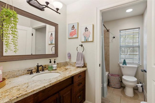 This bathroom features a granite countertop with a single sink and dark fixtures. It has a large framed mirror, wall art, and a cozy shower area with a window for natural light. | Image 33